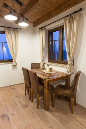 A dining area with a wooden table and chairs, a window with curtains, and a ceiling with exposed beams.
