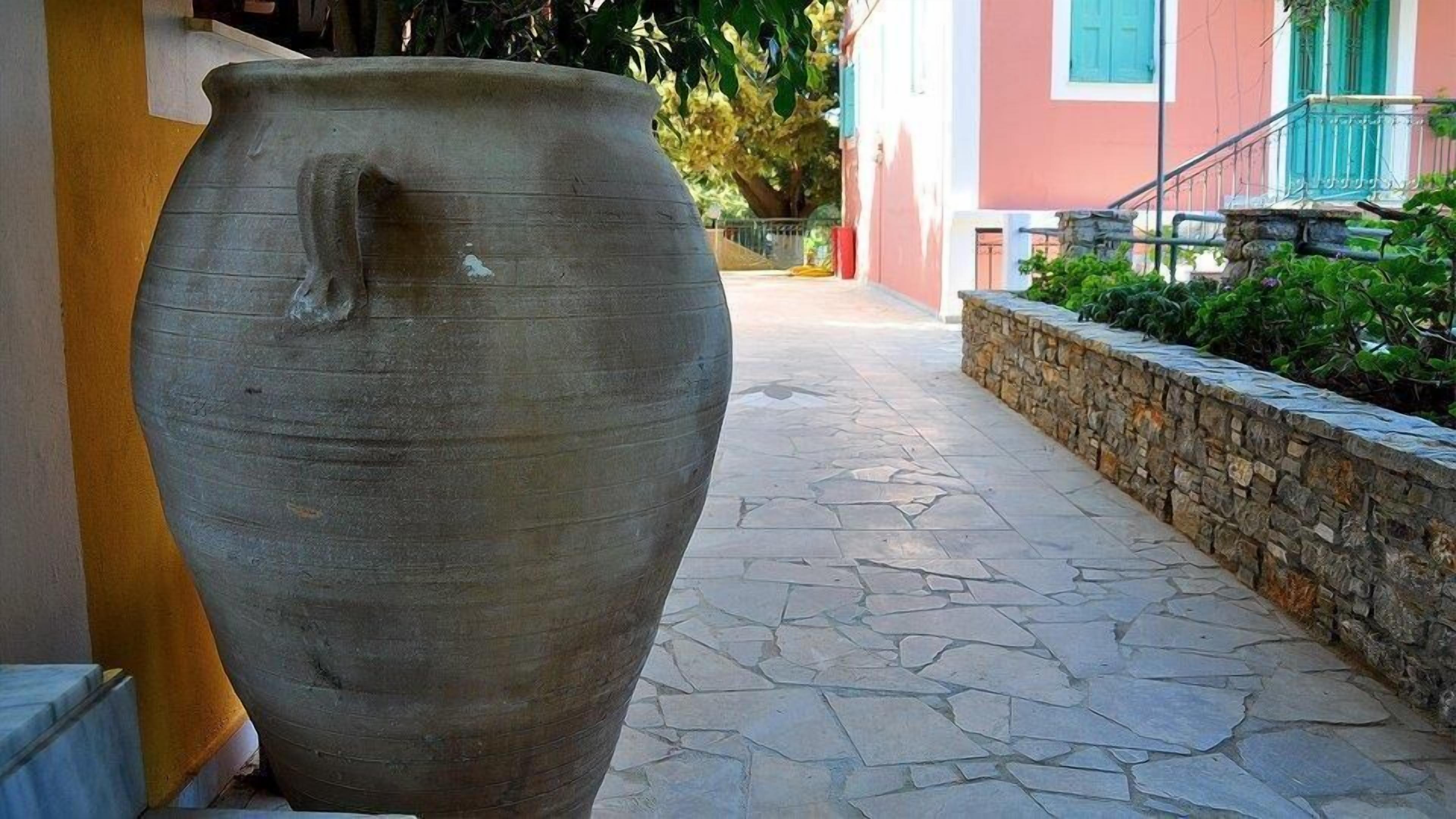 A large ceramic pot on a stone-paved path next to a building with a stone wall and greenery.
