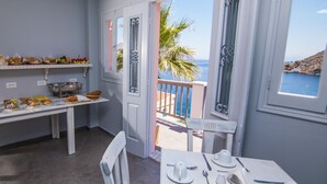 En matplats med ett bord dukat för fyra personer, en buffé med frukt och bakverk samt utsikt över havet genom den öppna dörren.