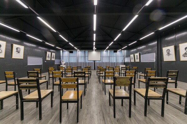 A conference room with rows of wooden chairs and tables, framed portraits on the walls, and a large screen at the front.