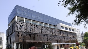 Ein modernes Gebäude mit einer markanten Fassade, die geometrische Ausschnitte aufweist, einem gut sichtbaren Eingang mit einem Schild und einem gepflegten Garten mit Steinen und Sträuchern.