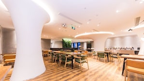 Ein modernes Restaurant mit Holztischen und grünen Stühlen, einer großen weißen Säule und einer grünen Wand mit einer Pflanze.
