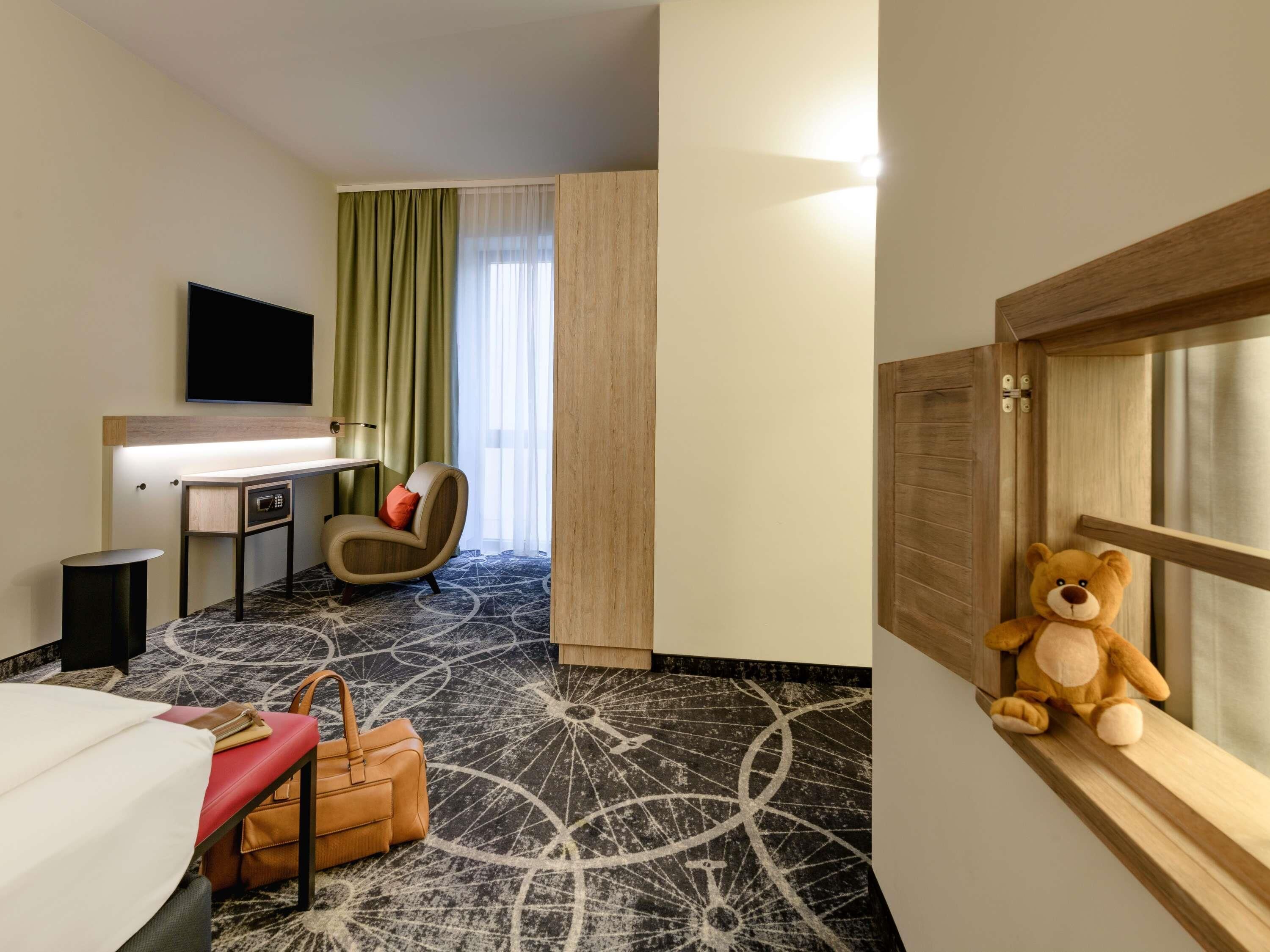 A hotel room with a bed, a desk, a chair, a teddy bear, and a window with curtains.