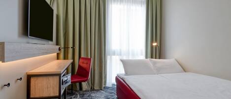 A hotel room with a bed, a desk, a chair, a TV, and a window with curtains.