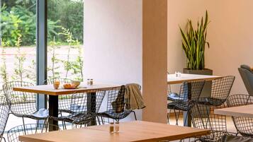 A modern cafe with metal wire chairs and wooden tables, large windows with a view of greenery, and a potted plant on the wall.