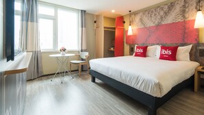 Une chambre d’hôtel avec un grand lit, une petite table, une chaise, une armoire et une fenêtre avec des rideaux.