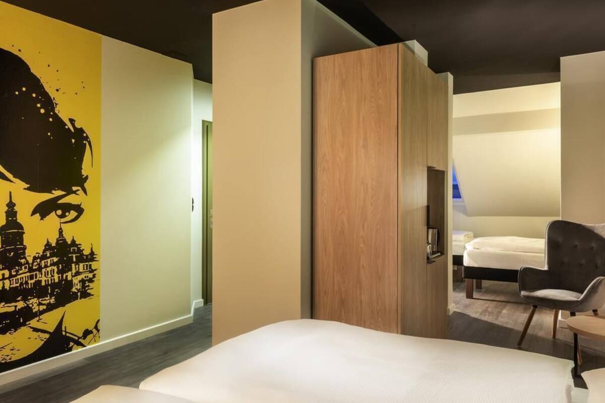 Une chambre d’hôtel moderne, équipée d’un lit, d’une chaise, d’une armoire en bois et d’un mur orné d’une fresque représentant un paysage urbain.