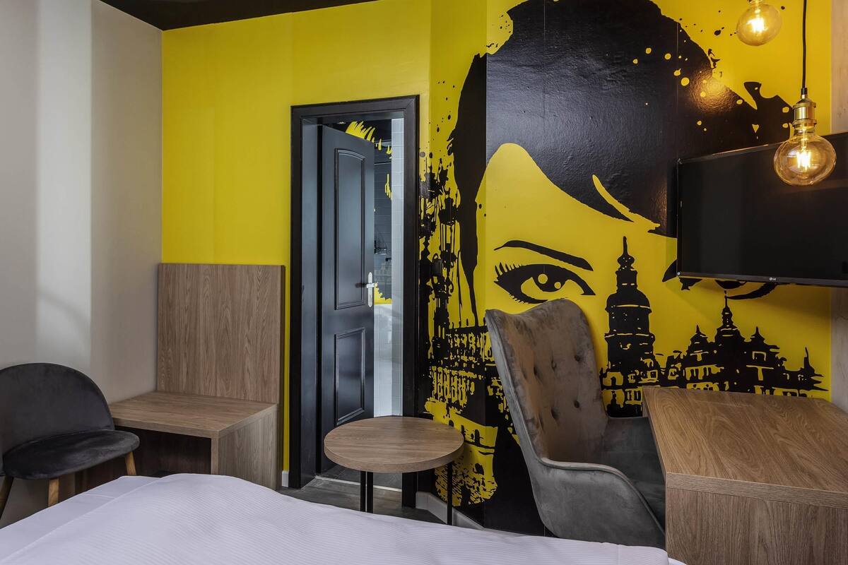 A hotel room with a yellow wall, a large mural, a bed, a desk, and two chairs.