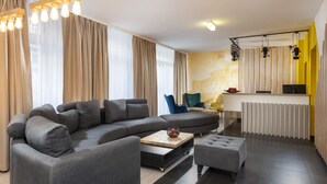 A modern living room with a sectional sofa, a coffee table, and a small ottoman. There is a kitchen area with a counter and a radiator. - ibis Styles Dresden Neustadt (Dresden)