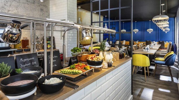 A buffet counter with various food items, a coffee machine, and a seating area with tables and chairs. - ibis Styles Dresden Neustadt (Dresden)