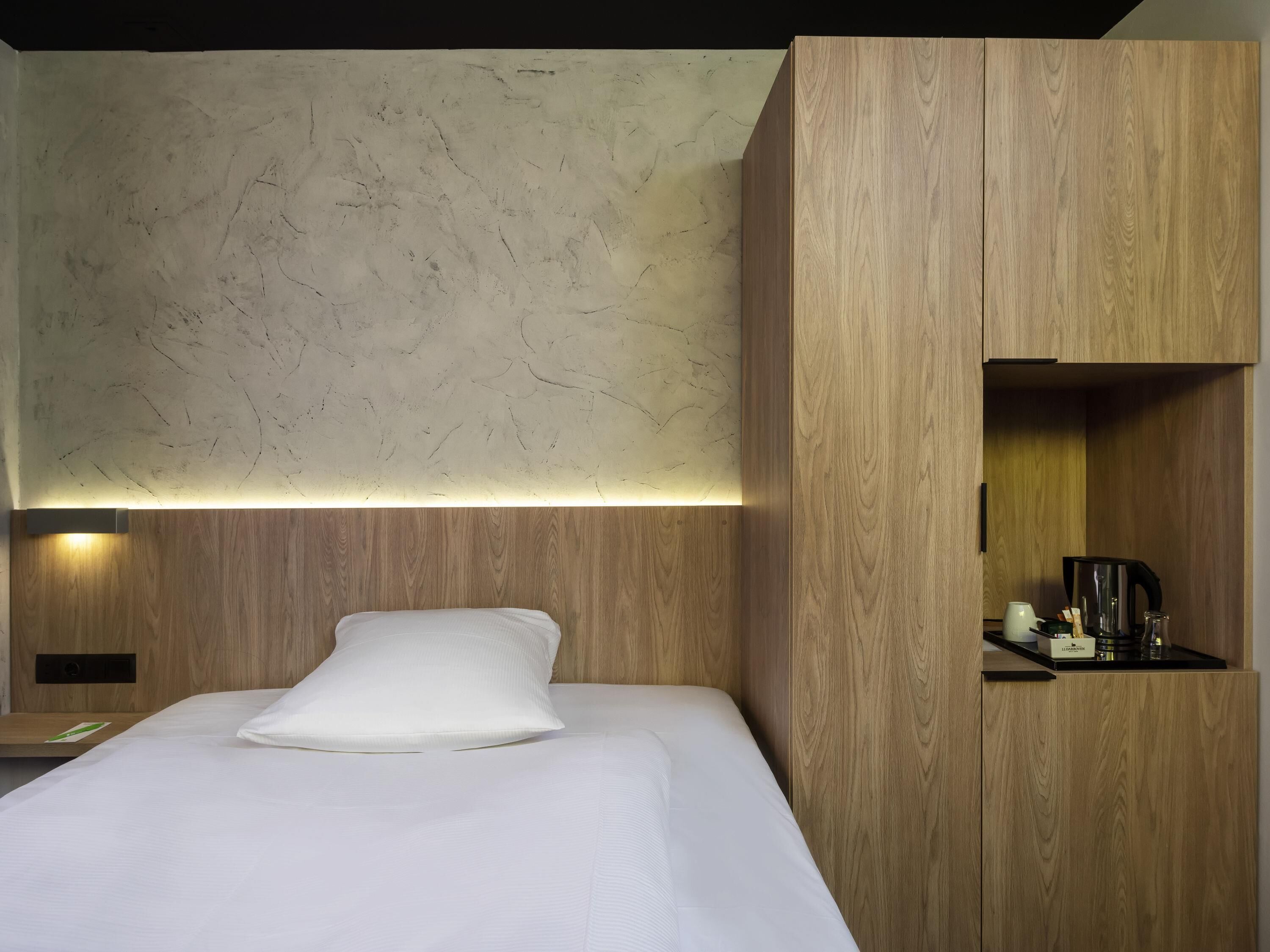 A hotel room with a bed, wooden headboard, a marble wall, and a shelf with a coffee maker.