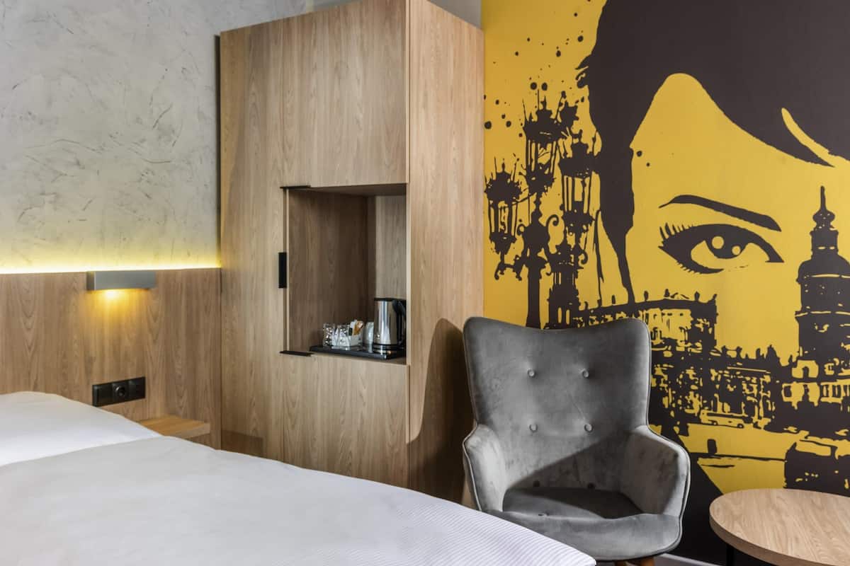 A hotel room with a bed, a grey armchair, a small table, a wooden wardrobe, and a wall with a black and yellow mural.
