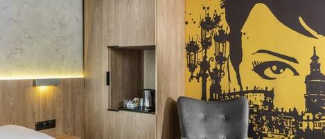 Habitación de hotel con cama, un sillón gris, una mesita, un armario de madera y un mural en la pared.