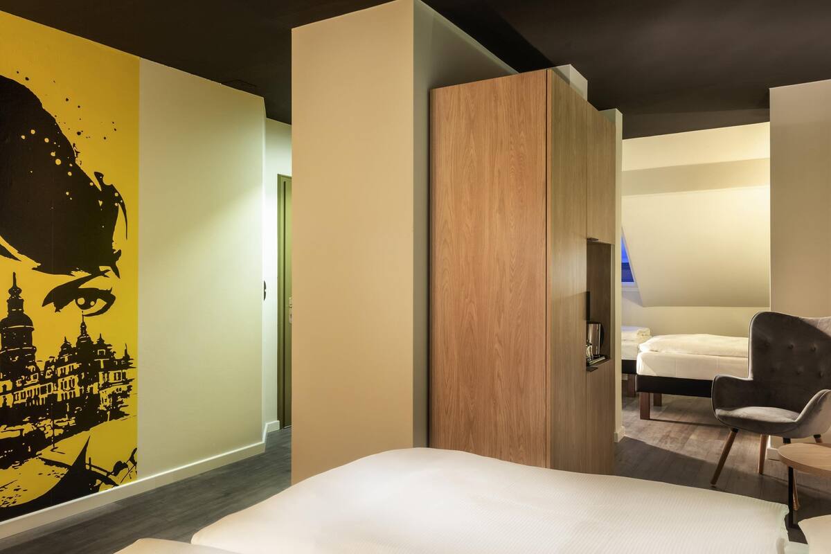 A hotel room with a bed, a chair, a wardrobe, and a wall with a cityscape mural.