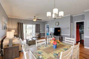 A spacious living room with a dining area, a ceiling fan, a flat-screen TV, and a kitchen in the background.