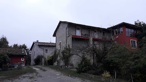 A rustic stone building with a red roof, a small balcony, and a garden area with trees and shrubs. - Cascina Binelli (Dogliani)