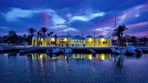 Un puerto deportivo con barcos amarrados, un edificio amarillo con restaurante y palmeras.