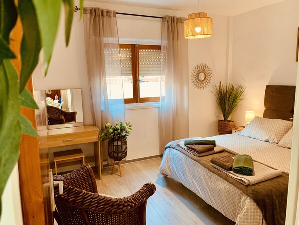 A bedroom with a bed, a desk, a chair, a window with curtains, a hanging light fixture, and a wall decoration.