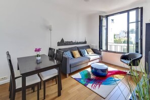 A modern living room with a sofa, a dining table, and chairs. There is a large window providing a view of the city. - Appartement Garros - Welkeys (Boulogne-Billancourt)
