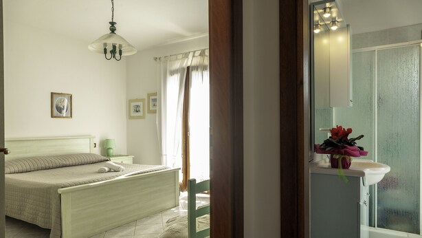 A bedroom with a bed, a chair, a nightstand, a lamp, and a bathroom with a sink and a mirror. - Aria Di Vacanza - Mirtillo COD 9 (Olbia)