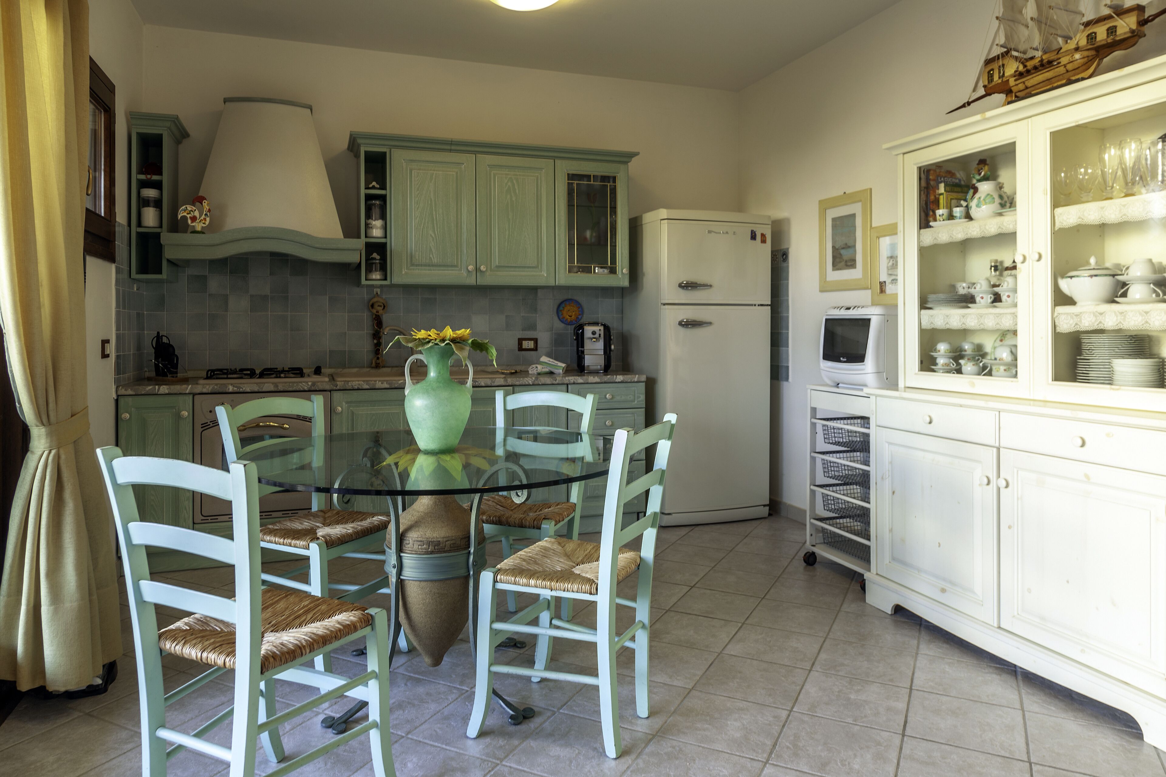 A kitchen with a dining table, chairs, a refrigerator, and a cabinet with dishes.