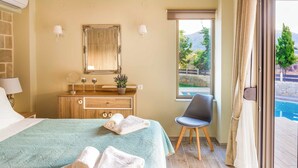 A bedroom with a bed, a wooden dresser, a mirror, and a window with a view of mountains and a pool. - Villa Elenitsa (Kissamos)