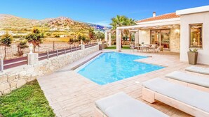 A pool area with a stone wall, lounge chairs, and a view of hills. - Villa Elenitsa (Kissamos)
