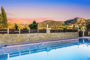 A swimming pool with stone walls and a wooden fence, set against a backdrop of hills and a sunset sky.
