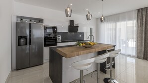Une cuisine moderne dotée d’un îlot central, d’appareils électroménagers en acier inoxydable, de luminaires suspendus et offrant une vue sur l’espace de vie.