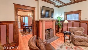 A living room with a fireplace, a television mounted above it, a dining area in the background, and a patterned rug on the floor.