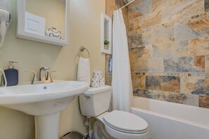 Casa de banho com lavatório branco, sanita e banheira. Tem cortina de chuveiro, um porta-toalhas e dispensador de sabonete.