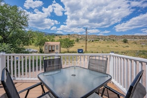 A deck with a glass-top table and chairs, overlooking a rural landscape with a small house and mountains in the distance. - 2BR Aptmt Parks Private Deck, Mountain Views! (Colorado Springs)