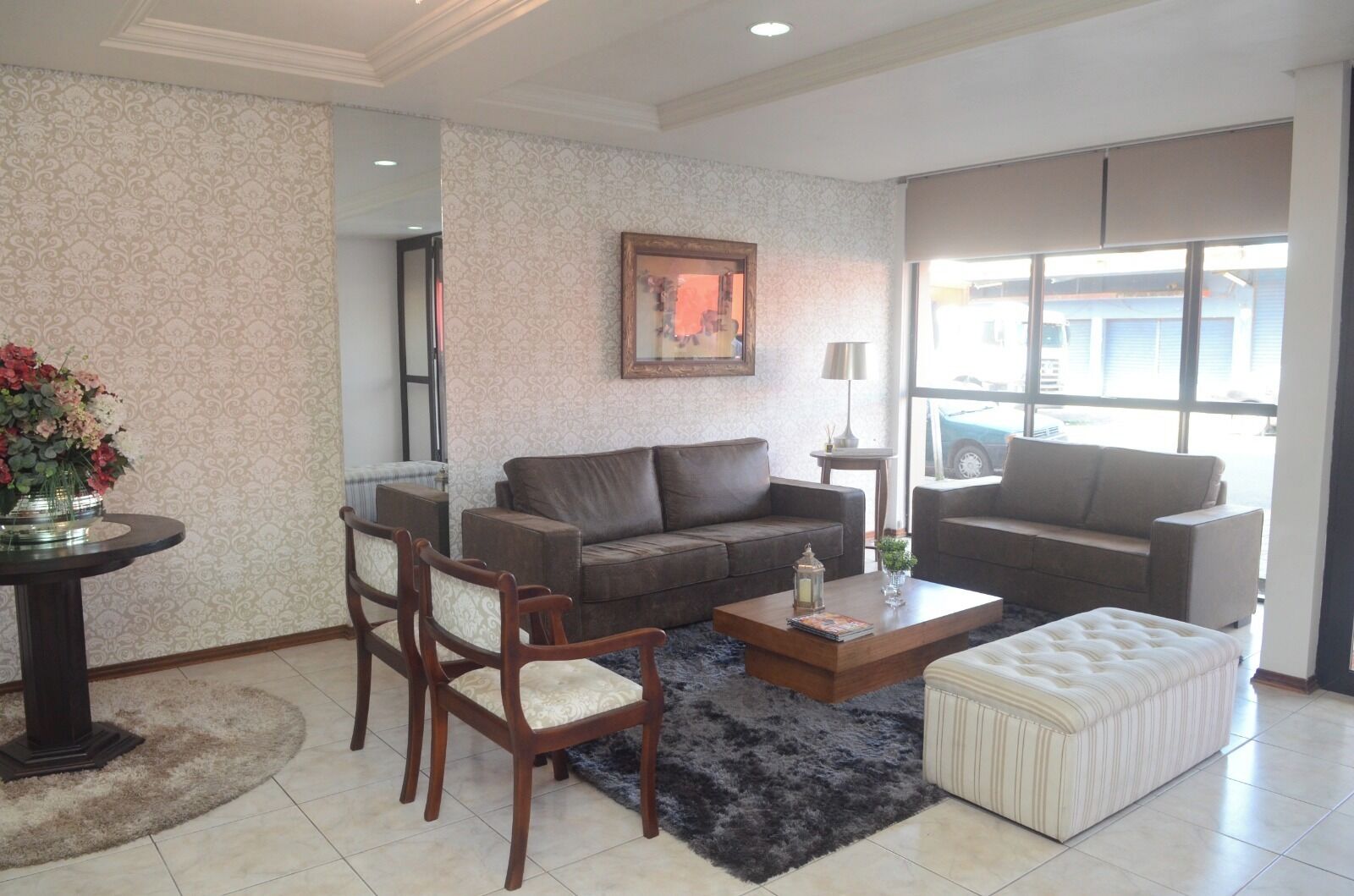 A living room with a brown sofa, a matching armchair, a coffee table, and a striped ottoman. A painting is on the wall, and there's a window with a view of the outside.