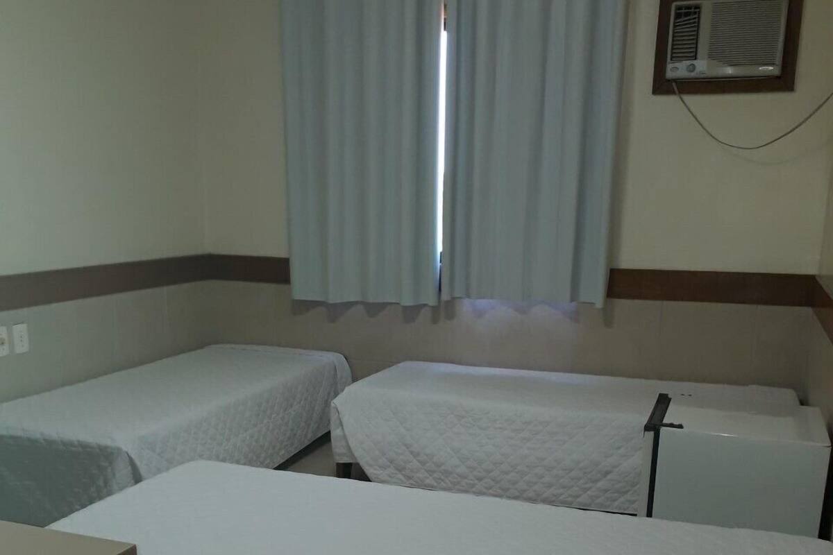 Una habitación con tres camas individuales, un escritorio pequeño, una ventana con cortinas y un equipo de aire acondicionado instalado en la pared.