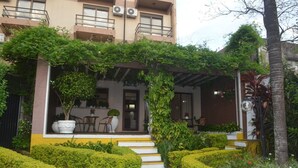 Un edificio con entrada cubierta, escalones que conducen a la puerta y un jardín con arbustos cuidados y un sendero.