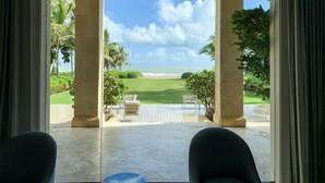 A view from a room with a large window overlooking a beach, palm trees, and a clear sky. - Luxurious Four Seasons PR Bahia Beach Villa; Golf, Pool, Tennis & Golf Cart (Río Grande)