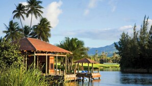 A wooden structure with a tiled roof over water, surrounded by palm trees and a golf course in the background. - Luxurious Four Seasons PR Bahia Beach Villa; Golf, Pool, Tennis & Golf Cart (Río Grande)
