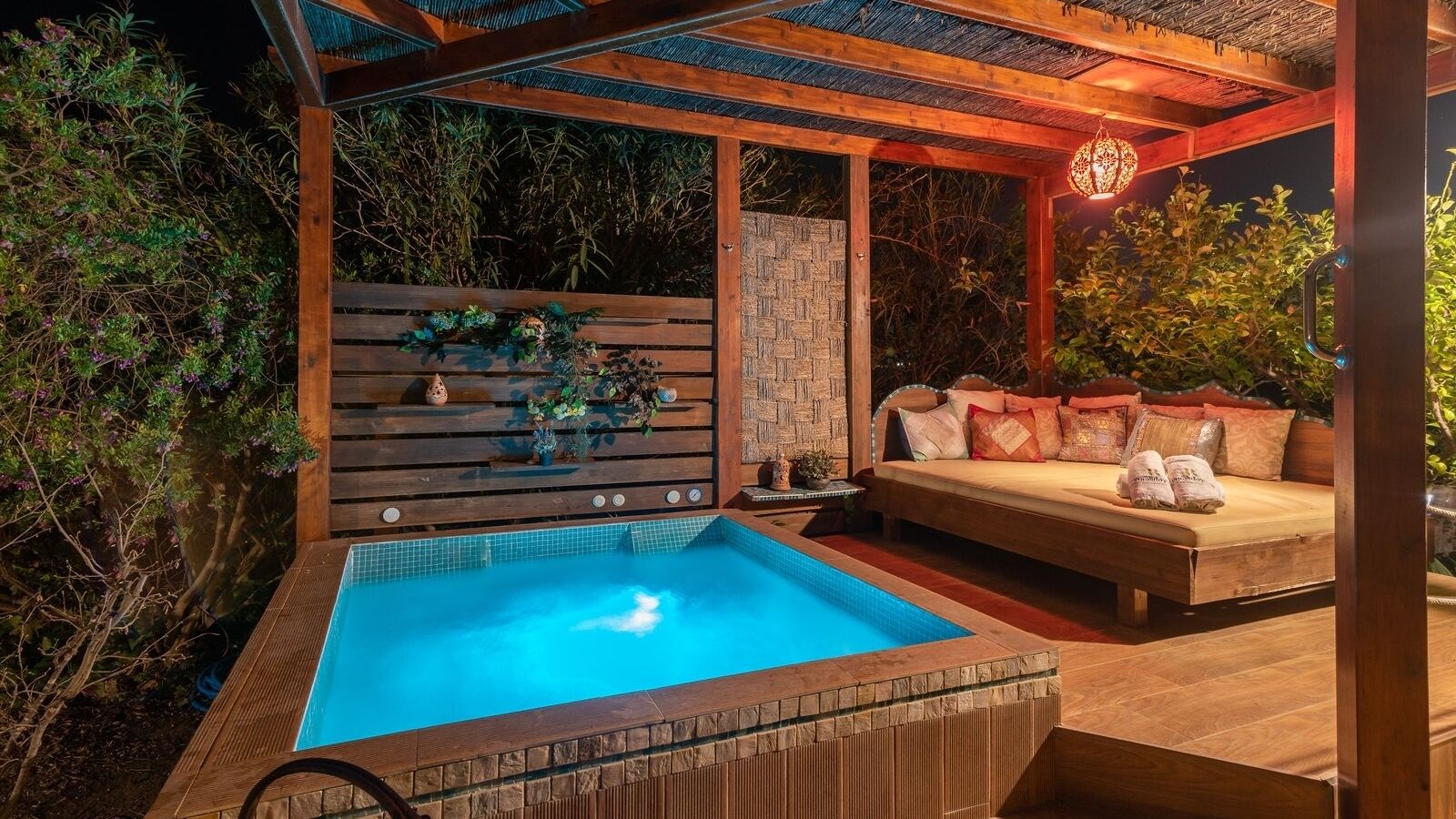 Een jacuzzi met een houten terras, een zithoek met kussens en een decoratieve muur met planten.