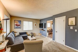 A living room with a gray sofa, a striped armchair, a coffee table, and a TV mounted on the wall. There is a window with blinds, a framed picture on the wall, and a door leading to another room. - 2BR Hikers Dream Red Rocks Dog-friendly! (Colorado Springs)