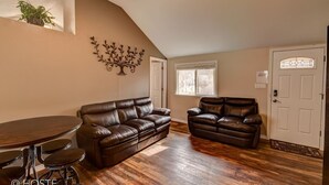 Un salon comprenant un canapé en cuir marron, un fauteuil en cuir marron et une table à manger en bois avec quatre chaises. Un élément décoratif orne le mur au-dessus du canapé, et une fenêtre avec des stores se trouve sur le mur.