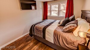 Une chambre à coucher avec un lit, un téléviseur fixé au mur, une fenêtre avec des rideaux et un plancher en bois.