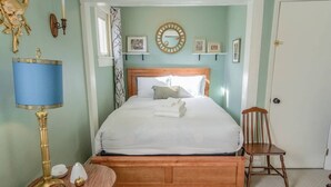 A neatly made bed with white linens, a wooden headboard, and a bedside table with a lamp. There are picture frames on the wall and a chair next to the bed. - Downtown 1900 Classic 3 Bedroom Value (Colorado Springs)