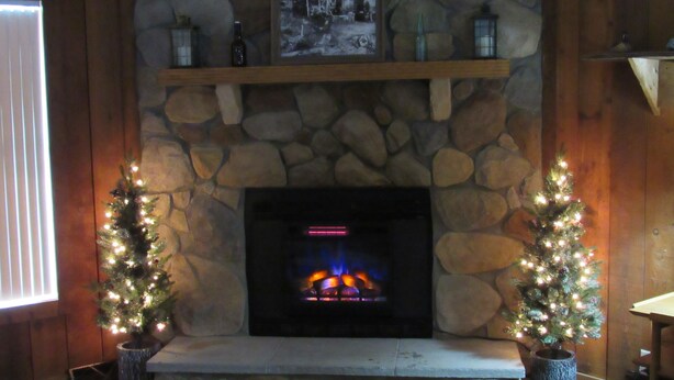 A stone fireplace with a lit fire, flanked by two decorated Christmas trees, set against a wooden wall with a stone shelf above. - Welcome to theThe Deer Drop Inn (Hulbert)
