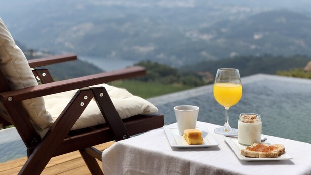 A dining table with a glass of orange juice, a cup, a jar of spread, and a slice of bread on a white tablecloth. - BHDouro (Baiao)