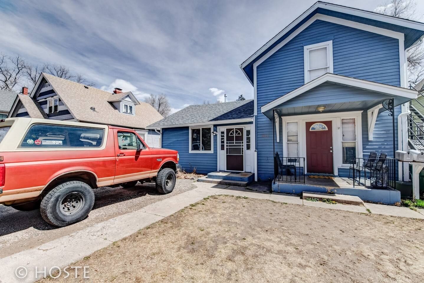 En röd pickup-truck parkerad framför ett blått hus med en veranda och en röd dörr.