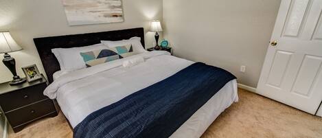 Una cama bien hecha con colcha blanca y azul oscuro, dos mesitas de noche con lámparas, un cuadro enmarco y un cuadro colgado en la pared.