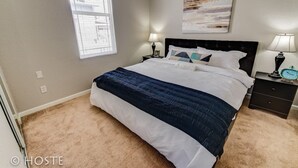 A bedroom with a large bed, two bedside tables with lamps, a wall-mounted painting, and a window with blinds.