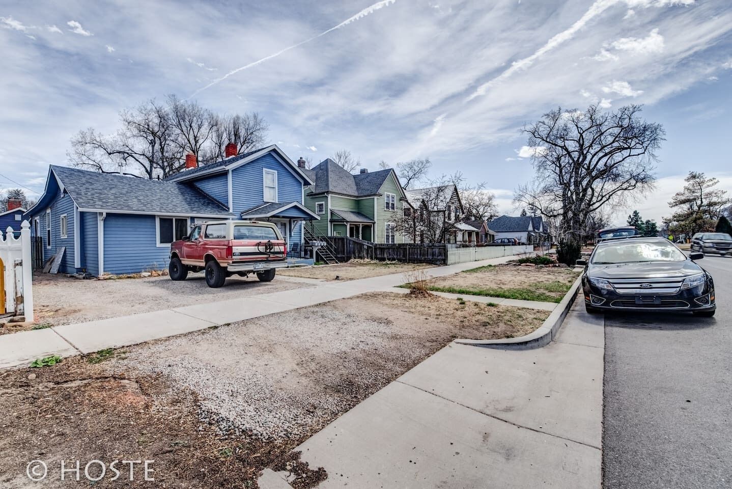 A residential street with houses, a red truck, and a black car parked on the side.