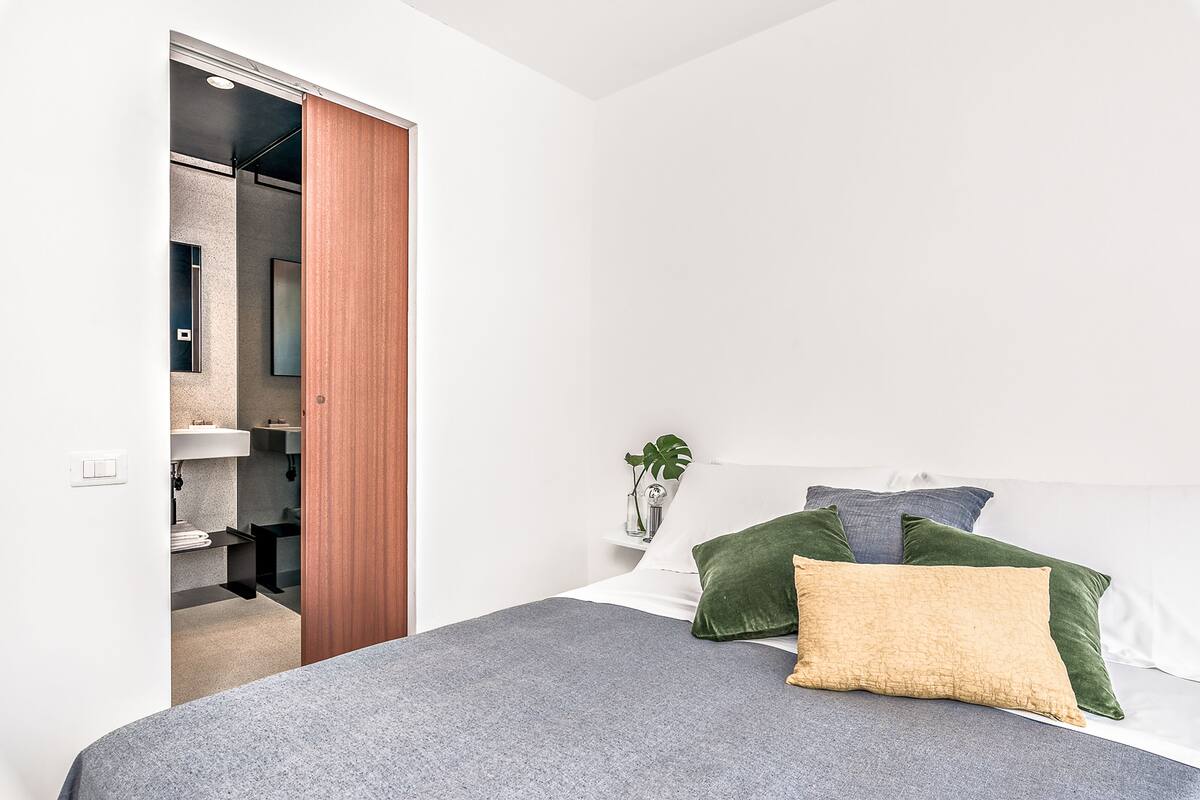 Ein Schlafzimmer mit Bett, Kissen, einem Nachttisch, einer Pflanze und einem durchgehenden Tür sichtbar erkennbaren Badezimmer.