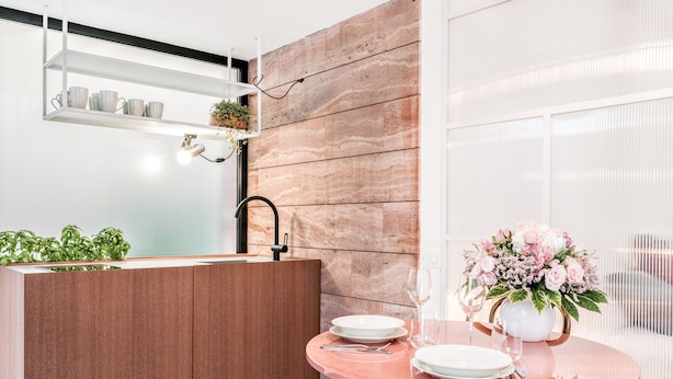 Un comedor con una mesa redonda, sillas y un mantel rosa. Se ve un aparador de madera con un estante que contiene tazas y una planta. Un jarrón con flores está sobre la mesa.